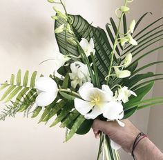 a hand holding a bouquet of white flowers and greenery in front of a wall