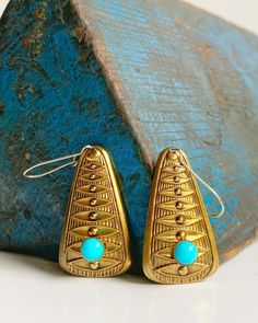 pair of gold earrings with turquoise stones on white table next to blue stone wall, closeup