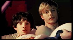 two young men laying in bed with one looking at the camera and the other staring