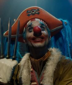 a clown dressed as a pirate with blue hair and red nose holding two knives in his hand