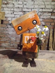 a wooden robot with flowers in it's hand standing next to a brick wall