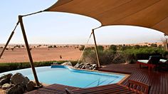 an outdoor swimming pool surrounded by wooden deck chairs and umbrellas with desert scenery in the background