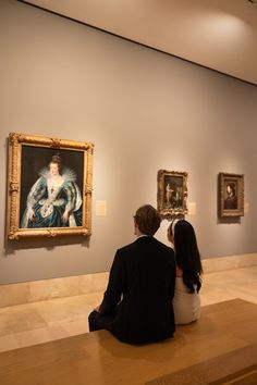 two people are sitting on a bench looking at paintings in an art museum, one is taking a photo