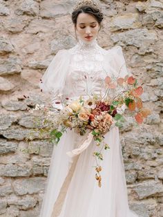 a woman wearing a wedding dress holding a bouquet in front of a stone wall with flowers on it