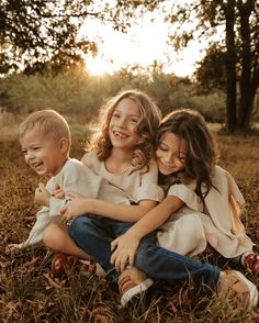 three young children sitting on the ground in front of trees and grass, smiling at the camera