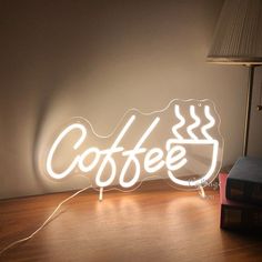 a neon coffee sign sitting on top of a wooden table next to a book and lamp