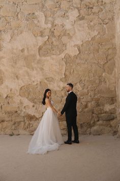 a bride and groom holding hands in front of a stone wall