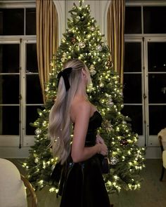 a woman with long blonde hair standing in front of a christmas tree wearing a black dress