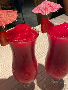 two glasses filled with red liquid and strawberries on top of a table next to an umbrella