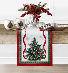 a christmas tree table runner with red berries and evergreens on it, next to two mugs