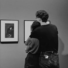 two people are looking at pictures on the wall in an art gallery, one is hugging the other's back