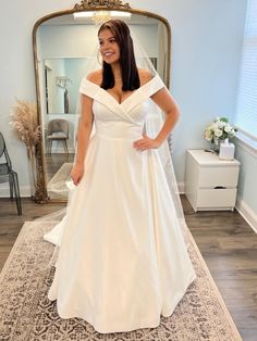 a woman standing in front of a mirror wearing a white wedding dress with off the shoulder sleeves