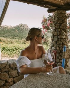 a woman sitting at a table with a glass of wine in her hand and mountains behind her