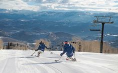 two people on skis going down a snowy hill with mountains in the back ground