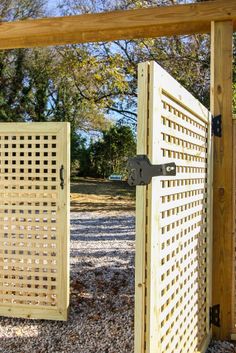 an open wooden gate in the middle of a gravel area with trees and bushes behind it