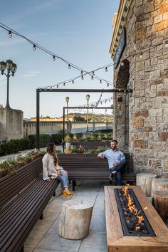 two people sitting on benches next to an outdoor fire pit with lights strung over it