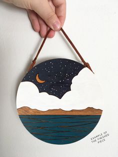 a hand is holding a wooden plaque with a night sky and stars above the water