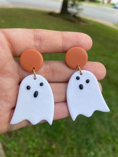 two white ghost earrings with black eyes and orange discs on their earwires are held in the palm of someone's hand