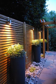 some plants are in large black pots on the side of a wooden fence at night