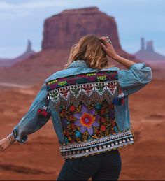 a woman wearing a jean jacket with an embroidered flower on the back, standing in front of a desert landscape