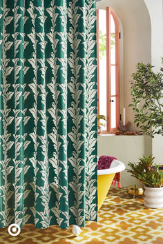 a green shower curtain with white leaves on it in front of a bathtub and potted plant