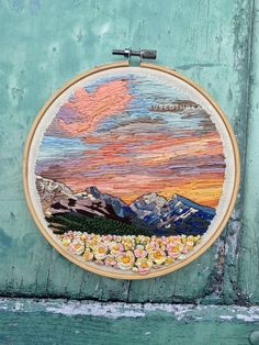 an embroidery project with mountains and flowers on the front is hanging on a green wall