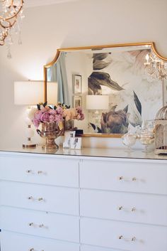 a white dresser topped with lots of drawers under a large framed mirror next to a chandelier