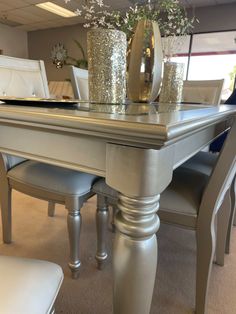 a dining room table with silver chairs and vases on top of it, in front of a window