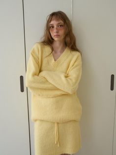 a young woman in a yellow sweater leaning against a white wall with her arms crossed