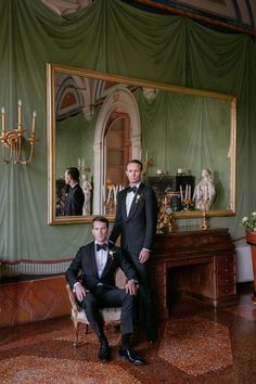 two men in tuxedos are posing for the camera, one is sitting on a chair