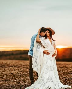 a pregnant woman in a white dress and cowboy hat hugs her husband as the sun sets