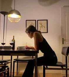 a woman sitting at a table writing on a piece of paper with a bottle of wine in the background