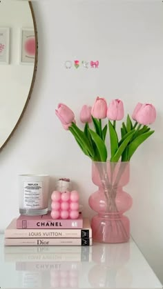 pink flowers are in a vase next to two books on a white table with a mirror