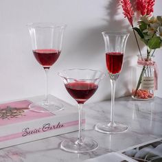 two glasses of red wine sit on a table next to a book and vase with flowers