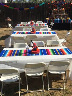 the table is set up with white chairs and colorful cloths on it for an outdoor party