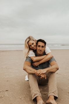 a man and woman sitting on the sand at the beach, hugging each other with an overcast sky in the background