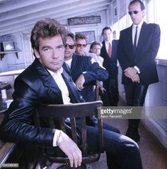 men in suits and ties sitting on chairs with one man looking at the camera behind them