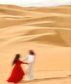 two people standing in the desert with sand dunes behind them and one person wearing a red dress