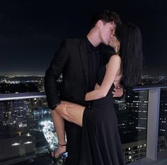 a man and woman kissing on top of a building at night with city lights in the background