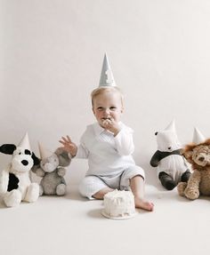 a baby sitting on the floor surrounded by stuffed animals wearing party hats and eating cake