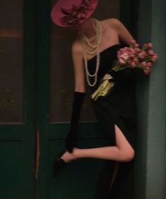 a woman in a black dress and pink hat leaning against a green door with flowers