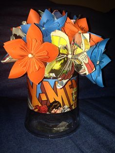 a glass vase filled with paper flowers on top of a blue cloth covered tablecloth