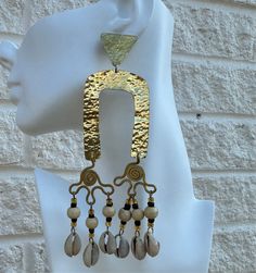 The Zanele Hammered Brass Cowry Earrings feature a striking hammered brass arch with intricate swirl designs and dangling beads, finished with elegant cowry shells. These earrings exude a blend of traditional charm and contemporary style, making them a unique statement piece for any occasion. Measures length 5.5 inches Gold Bohemian Chandelier Earrings For The Beach, Bohemian Gold Chandelier Earrings For Beach, Artisan Gold Earrings For Beach, Festival Brass Earrings With Gold Beads, Brass Earrings With Gold Beads For Festivals, Cowry Shell, Hammered Brass, Swirl Design, Ring Bracelet