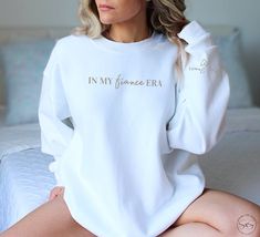 Elevate your bridal style with our 'In My Fiance Era' sweatshirt - a chic blend of modern elegance and timeless tradition. Customize with your wedding date or last name on the sleeve accompanied by a sweet heart detail.  Makes a great gift for those newly engaged or a thoughtful addition to any wedding shower! The ideal gift for brides-to-be, this sweatshirt is a delightful way to share in the joy of their journey. Elevate your bridal style with this must-have! Taking care of your sweatshirt to ensure it stays cozy and stylish for the long run is easy. For best results, machine wash it inside out with like colors in cold water. Use a mild detergent and avoid bleach. Tumble dry on low heat, or hang it to air dry for optimal longevity. If needed, iron it on low heat, but avoid ironing direct Mrs Sweatshirt, Womens Sweatshirts, Newly Engaged, Bride Gift, Bridal Shower Gift, Sweet Heart, Bridal Shower Gifts, Modern Elegance, Bride Gifts