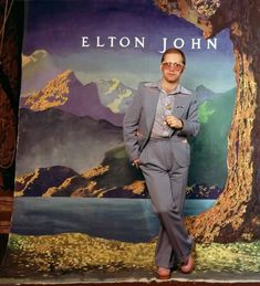 a man standing in front of a backdrop with a tree and mountains behind him wearing sunglasses