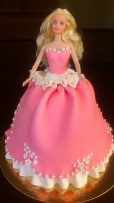 a barbie doll cake with pink dress and white flowers on the bottom, sitting on a gold plate