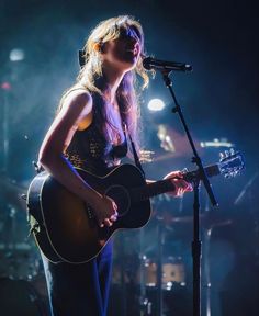 a woman holding a guitar while standing in front of a microphone on top of a stage