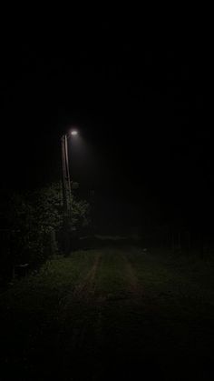 a dark road at night with a street light in the distance and trees on either side