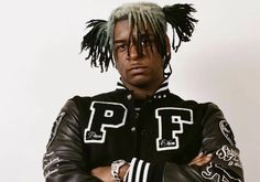 a young man with dreadlocks standing in front of a white wall wearing a black jacket