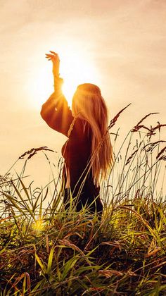 a woman is standing in tall grass with her arms up and the sun shining behind her